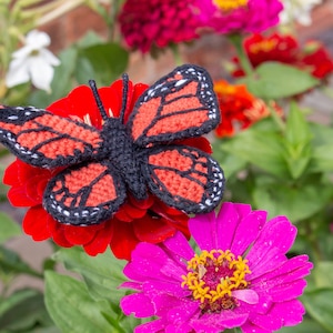 May include: A crocheted butterfly with black and orange wings is perched on a red zinnia flower. The butterfly is surrounded by other pink and red flowers.