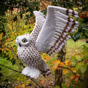 May include: A white crocheted snowy owl with brown accents on its wings, perched on a branch with autumn leaves in the background.