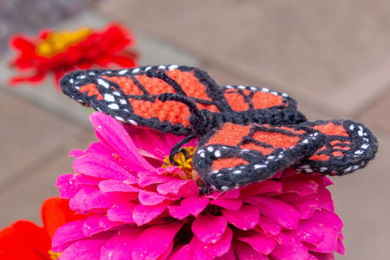 May include: A crocheted butterfly with black and orange wings is perched on a pink zinnia flower.
