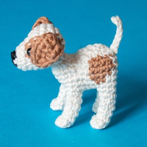 May include: A crocheted white dog with brown patches, standing on a blue background.