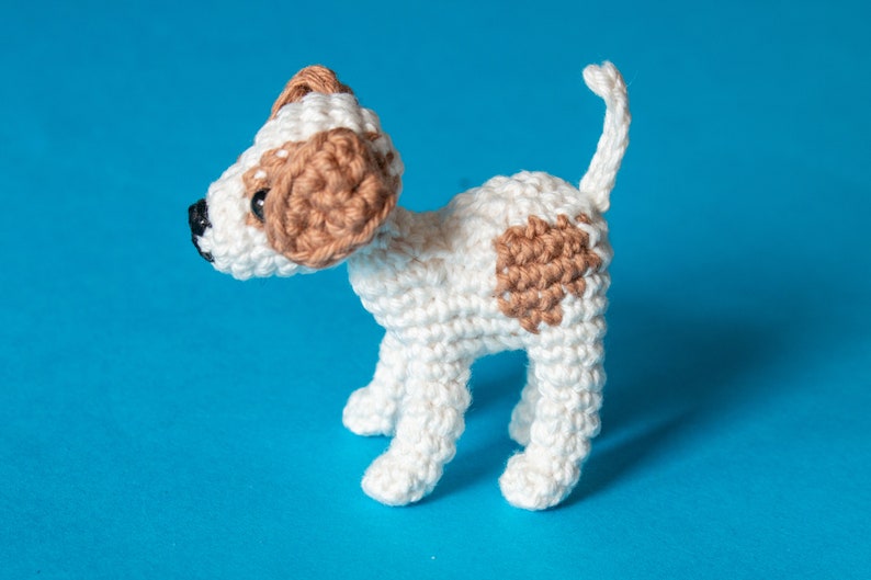 May include: A crocheted white dog with brown patches, standing on a blue background.