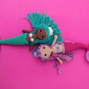 May include: Two crocheted mermaid dolls on a pink background. One mermaid has a green tail and brown skin, the other has a purple tail and light skin.