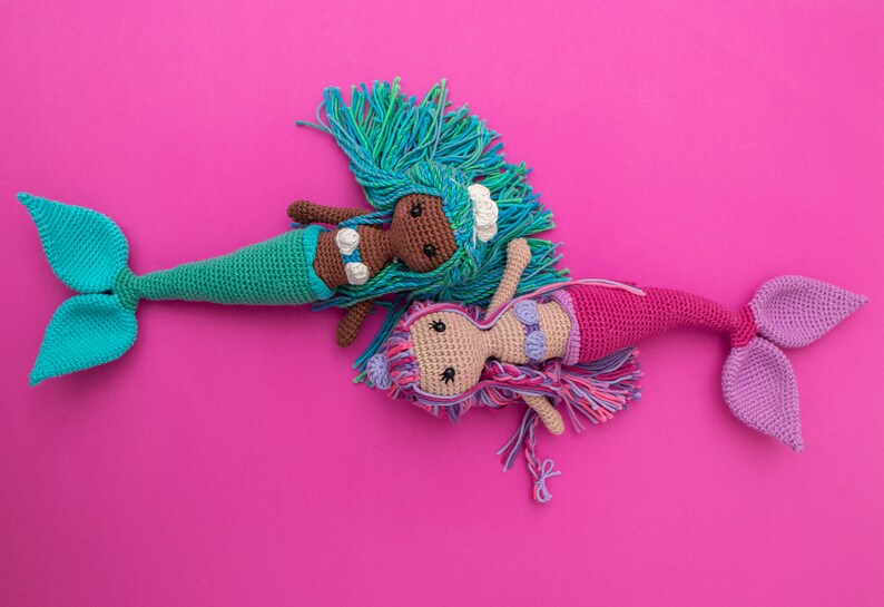 May include: Two crocheted mermaid dolls on a pink background. One mermaid has a green tail and brown skin, the other has a purple tail and light skin.
