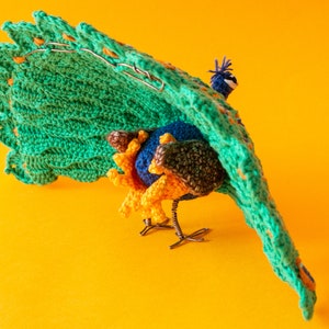K&ouml;nnte beinhalten: Eine handgefertigte Pfauenskulptur mit einer lebendigen Farbpalette. Der Pfau hat einen gro&szlig;en, gr&uuml;nen geh&auml;kelten Schwanz mit orangefarbenen Akzenten, einen blauen K&ouml;rper und gelbe und braune Details. Die Skulptur steht vor einem leuchtend gelben Hintergrund.