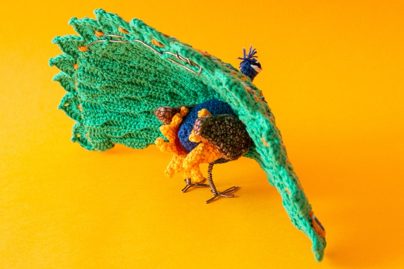 K&ouml;nnte beinhalten: Eine handgefertigte Pfauenskulptur mit einer lebendigen Farbpalette. Der Pfau hat einen gro&szlig;en, gr&uuml;nen geh&auml;kelten Schwanz mit orangefarbenen Akzenten, einen blauen K&ouml;rper und gelbe und braune Details. Die Skulptur steht vor einem leuchtend gelben Hintergrund.