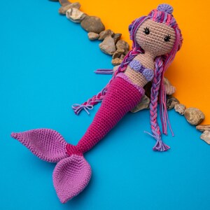 May include: A crocheted mermaid doll with pink and purple yarn. The doll has a purple tail and a pink top. The doll is lying on a blue and orange background.