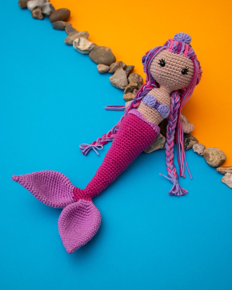 May include: A crocheted mermaid doll with pink and purple yarn. The doll has a purple tail and a pink top. The doll is lying on a blue and orange background.