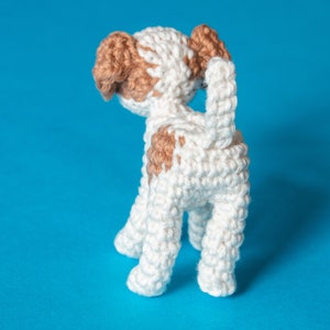 May include: A white crocheted dog with brown patches, standing on a blue background. The dog is facing away from the camera, with its tail curled up.