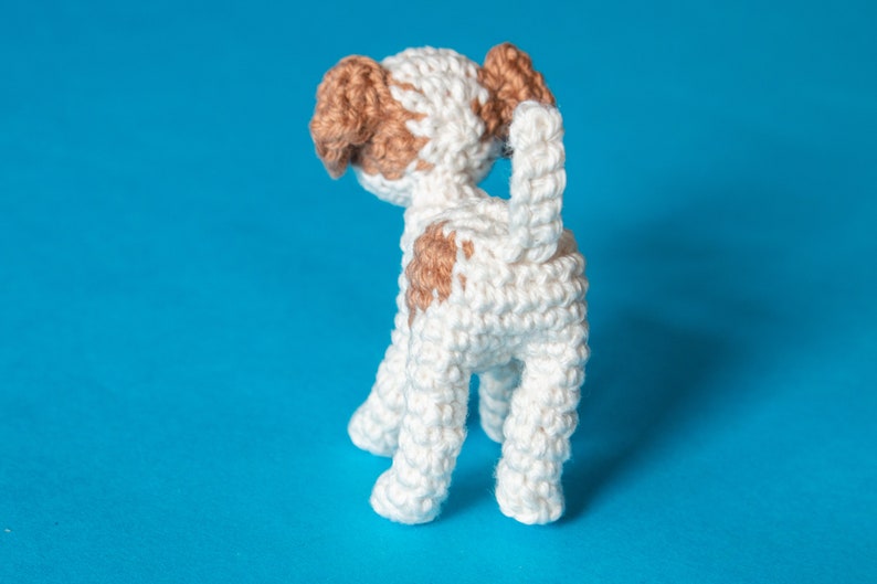 May include: A white crocheted dog with brown patches, standing on a blue background. The dog is facing away from the camera, with its tail curled up.
