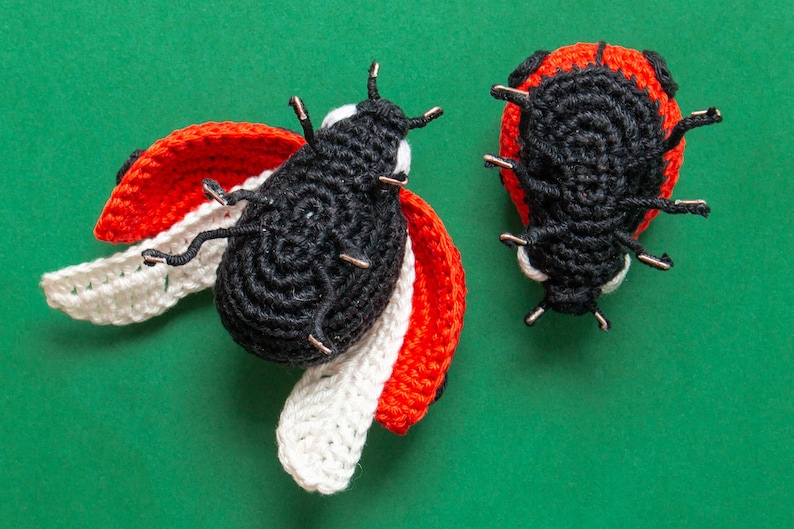 Puede incluir: Dos mariquitas hechas a mano con ganchillo sobre un fondo verde. Una mariquita tiene las alas abiertas con detalles rojos y blancos. La otra mariquita est&aacute; cerrada. Ambas mariquitas son negras con detalles rojos y tienen antenas de alambre.
