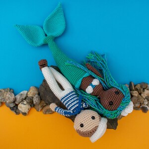 May include: A crocheted mermaid doll with blue and green hair, wearing a white seashell bra, lies on a rock with a crocheted sailor doll. The sailor doll is wearing a blue and white striped shirt and a white hat.
