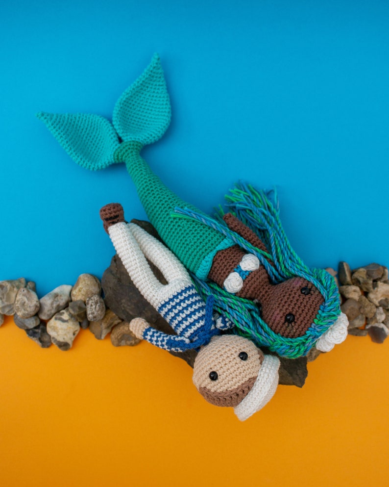 May include: A crocheted mermaid doll with blue and green hair, wearing a white seashell bra, lies on a rock with a crocheted sailor doll. The sailor doll is wearing a blue and white striped shirt and a white hat.