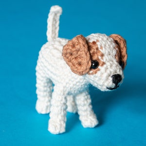 May include: A crocheted white and brown dog toy with black eyes. The dog is standing on a blue background.