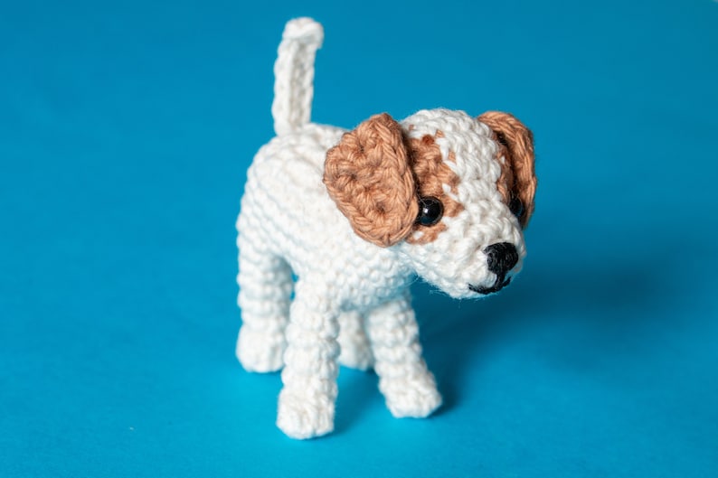May include: A crocheted white and brown dog toy with black eyes. The dog is standing on a blue background.