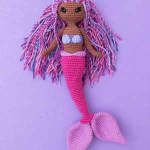 May include: A crocheted mermaid doll with pink and purple hair, a pink tail, and a purple seashell bra. The doll is sitting on a purple background.