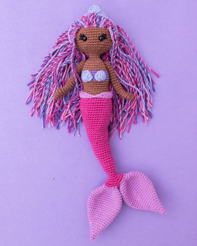 May include: A crocheted mermaid doll with pink and purple hair, a pink tail, and a purple seashell bra. The doll is sitting on a purple background.