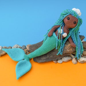 May include: A crocheted mermaid doll with brown skin, turquoise hair, and a turquoise tail. The doll is sitting on a rock with a blue background.