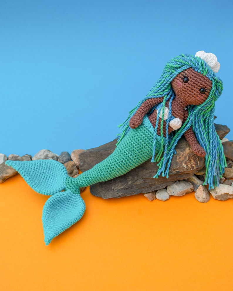 May include: A crocheted mermaid doll with brown skin, turquoise hair, and a turquoise tail. The doll is sitting on a rock with a blue background.