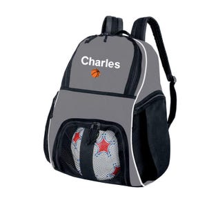 May include: A grey backpack with black accents, featuring the name Charles printed in white above an orange basketball icon. The front includes a black mesh pocket containing a white ball with red star patterns and black trim.