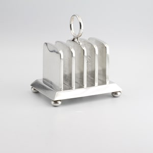 May include: A silver toast rack with four compartments and a ring handle. The rack sits on a rectangular base with four small, rounded feet. The design is simple and elegant.