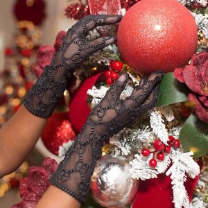May include: A pair of black lace gloves with a repeating pattern of interlocking Gs. The gloves are being used to decorate a Christmas tree with red ornaments and white flocked branches.