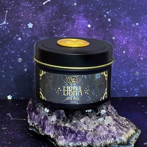 May include: A black candle tin with a gold lid and label. The label says "Libra Love Spell" and features a gold star design. The candle is sitting on a purple amethyst crystal.