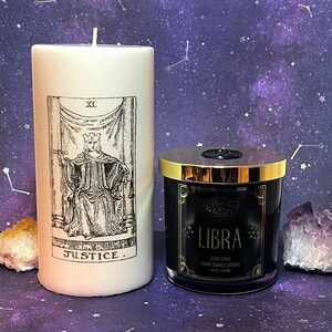 May include: Two candles, one white with a black and white illustration of the Justice tarot card and the text "JUSTICE" and one black with gold lettering that says "LIBRA LOVE SPELL CLEAR QUARTZ CRYSTAL 10 oz | 140 hrs".
