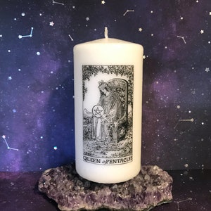 May include: White pillar candle with a black and white image of the Queen of Pentacles tarot card. The candle is sitting on a purple amethyst crystal cluster.