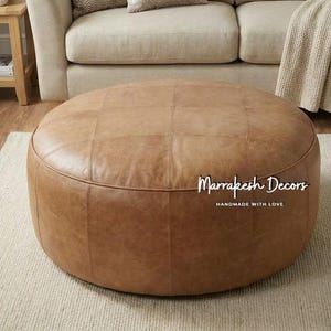May include: A large, round, brown leather ottoman with a stitched, grid-like pattern. The ottoman is centered on a light-colored rug, with a beige sofa and wooden side table in the background. The text "Marrakesh Decors HANDMADE WITH LOVE" is visible.