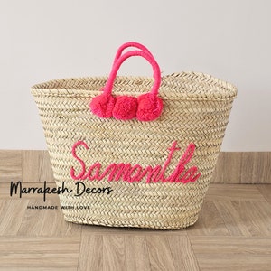 May include: A large straw tote bag with pink pom poms and the name "Samantha" embroidered in pink. The bag is made by Marrakech Decors and says "Handmade with Love".
