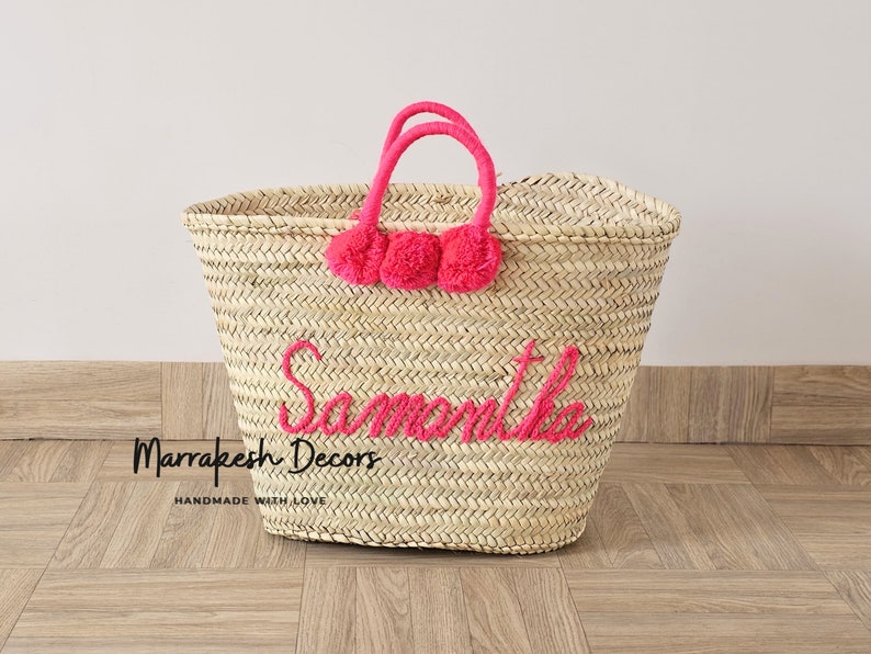May include: A large straw tote bag with pink pom poms and the name "Samantha" embroidered in pink. The bag is made by Marrakech Decors and says "Handmade with Love".