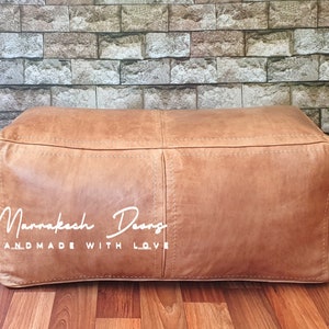 May include: A rectangular, light brown leather ottoman with visible stitching and a textured surface. The ottoman is placed on a wooden floor, with a brick-patterned wall in the background. The text "Mannakesh Design HANDMADE WITH LOVE" is visible on the ottoman.
