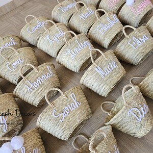 May include: A collection of personalized straw tote bags with white embroidered names. The bags are arranged in a grid pattern on a wooden surface. The bags are a natural straw color and have a woven texture.