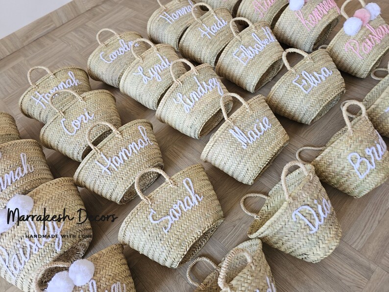 May include: A collection of personalized straw tote bags with white embroidered names. The bags are arranged in a grid pattern on a wooden surface. The bags are a natural straw color and have a woven texture.
