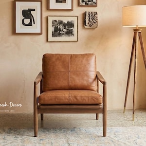 May include: A brown leather armchair with a wooden frame. The chair is in a room with a wooden floor lamp and framed artwork on the wall. The room has a neutral colour palette and a rug.