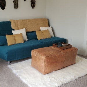 Puede incluir: Un sofá cama azul teal con una manta de tiro de color canela y almohadas. Un otomano de cuero marrón con una bandeja de madera en la parte superior se encuentra sobre una alfombra blanca y esponjosa.