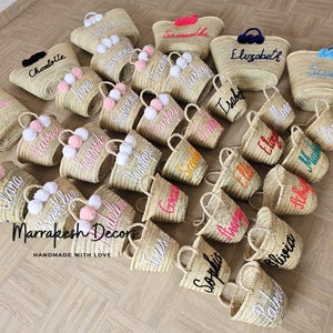 May include: A collection of straw tote bags with pom poms and personalized embroidery. The bags are in various colors and feature names like "Clara", "Charlotte", "Elizabeth", "Olivia", and "Sophia".