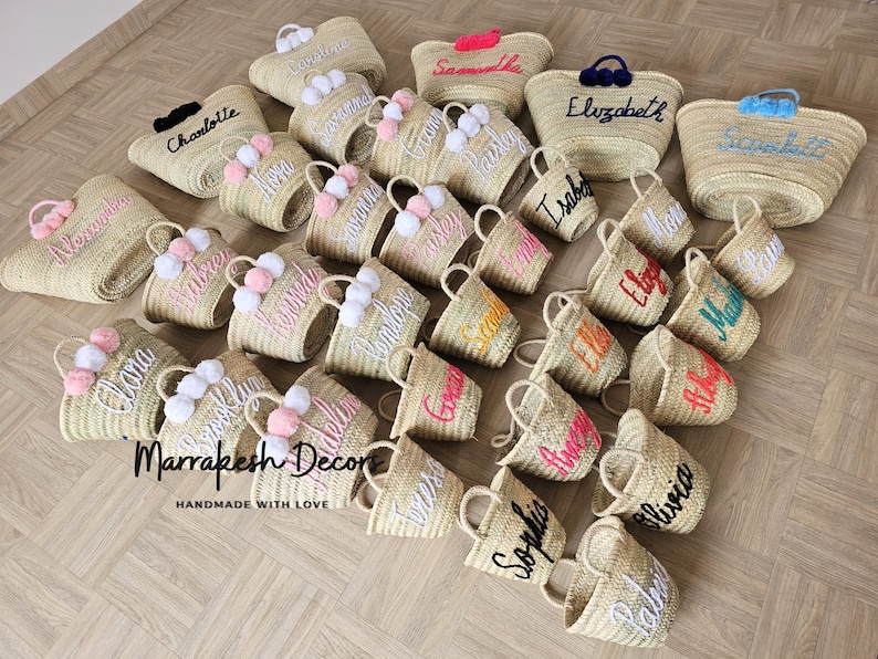 May include: A collection of straw tote bags with pom poms and personalized embroidery. The bags are in various colors and feature names like "Clara", "Charlotte", "Elizabeth", "Olivia", and "Sophia".