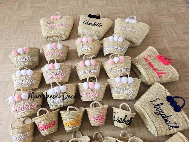 May include: A collection of straw tote bags with personalized embroidery and pom-poms. The bags are in various sizes and colors, with names like "Clara", "Savannah", "Paisley", and "Elizabeth" embroidered on them.