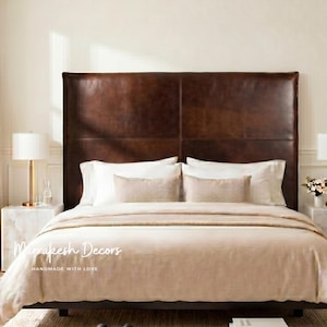 May include: A bed with a large, dark brown leather headboard. The bed is made with white pillows, beige bedding, and two matching beige bolster pillows. Two marble bedside tables with lamps flank the bed.