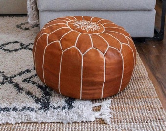 May include: A brown leather pouf with white stitching and a white embroidered design. The pouf is sitting on a rug with a white and gray pattern.