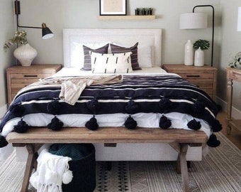 Könnte beinhalten: Ein weißes Bett mit schwarz-weiß gestreiftem Bettzeug mit schwarzen Bommeln. Das Bett hat ein Kopfteil aus Holz und eine Holzbank am Fußende des Bettes. Auf beiden Seiten des Bettes stehen zwei Nachttische mit Lampen.