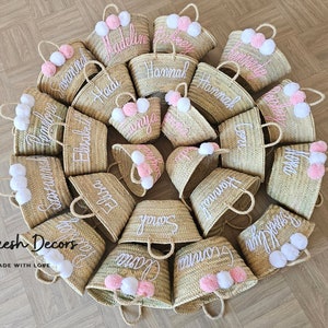 May include: A collection of 16 straw tote bags with personalized names embroidered in white thread. Each bag has a pink and white pom-pom attached to the handle. The bags are arranged in a circle on a wooden floor. The text "Marrakesh Decors Handmade with Love" is visible at the bottom of the image.