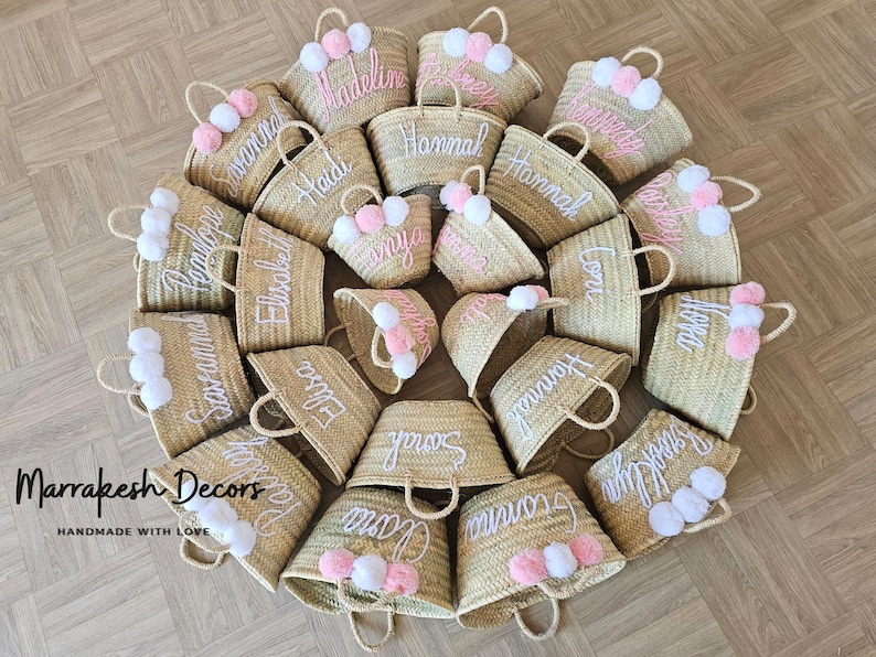 May include: A collection of 16 straw tote bags with personalized names embroidered in white thread. Each bag has a pink and white pom-pom attached to the handle. The bags are arranged in a circle on a wooden floor. The text "Marrakesh Decors Handmade with Love" is visible at the bottom of the image.