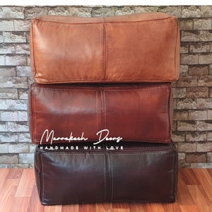 May include: Three stacked leather ottomans in shades of brown, from light tan to deep burgundy. Each ottoman is rectangular with visible stitching and a padded appearance. The text "Marrakech Doors" and "HANDMADE WITH LOVE" is visible on the middle ottoman.