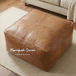 Puede incluir: Un otomán cuadrado de cuero marrón con detalles cosidos. El otomán está sobre una alfombra de color neutro. El texto "Marrakesh Decors" y "HANDMADE WITH LOVE" son visibles en la parte delantera del otomán.