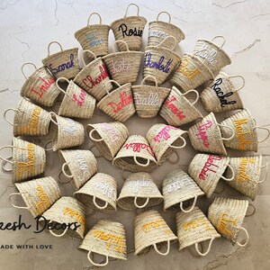 May include: A collection of 20 small, natural woven straw baskets with personalized names embroidered in various colors. The baskets are arranged in a circle on a light beige surface. The text "Marrakesh Decors" and "HANDMADE WITH LOVE" is visible at the bottom of the image.