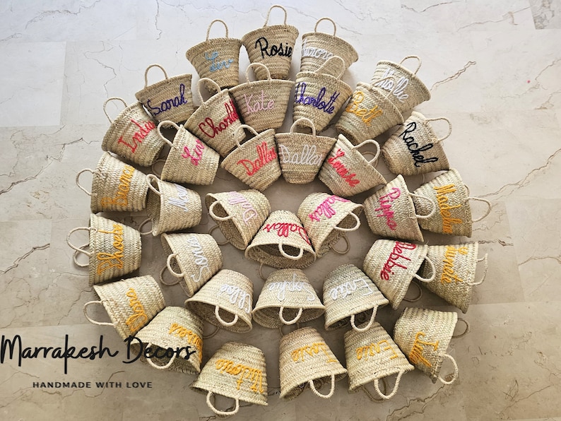 May include: A collection of 20 small, natural woven straw baskets with personalized names embroidered in various colors. The baskets are arranged in a circle on a light beige surface. The text "Marrakesh Decors" and "HANDMADE WITH LOVE" is visible at the bottom of the image.