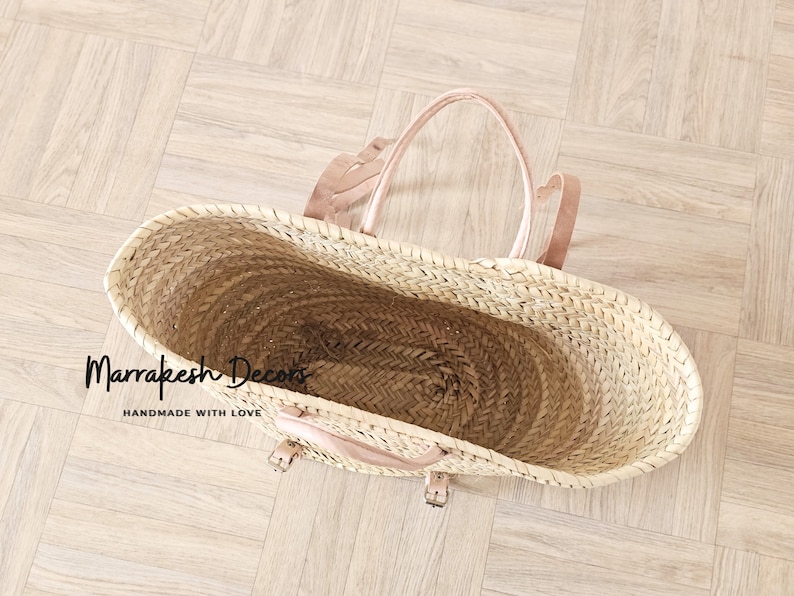 May include: A large, oval-shaped, woven straw tote bag with leather handles and buckles. The bag is empty and the inside is visible. The text "Marrakesh Decors Handmade with Love" is printed on the bottom of the bag.