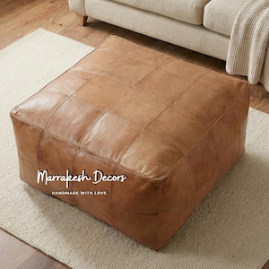 Peut inclure: Un pouf carré en cuir marron avec un motif cousu en forme de grille. Le pouf est posé sur un tapis crème, avec un canapé beige en arrière-plan. Le texte "Marrakesh Decors HANDMADE WITH LOVE" est visible sur le devant du pouf.